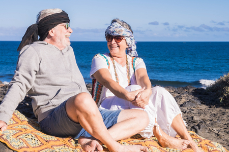 happy couple of senior people gentleman and lady sit down near the ocean with hippy colored clothes style. enjoy and have fun with alternative lifestyleの写真素材