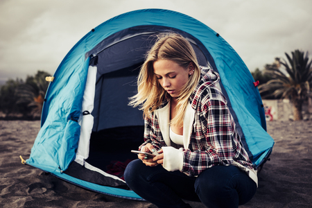 Always connected with technology blonde young woman stay ouside of her tent on the beach to enjoy freedom from work.の写真素材
