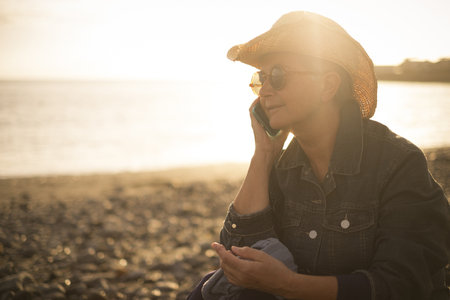 beautiful senior woman speak at the phone on the beach during a sunset. amazing golden warm backlight. outdoor lifestyle leisure concept working and living near the ocean. natural place to live greatの写真素材