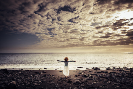 epic beautiful woman with amazing swirl hair open arms and hug the wonderful world outdoor at the beach with warm sunset and clouds in front of her.の写真素材