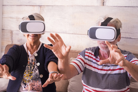 hands to protect while a caucasian man and woman aged 70 years old play at home with internet technology virtual reality goggles headset. smile and laugh together in a nice dayの写真素材