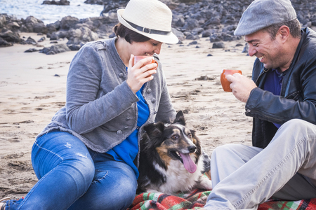 beautiful middle age couple in leisure on the beach in vacation summer time with a cute pet border collie. all together with a cup of coffee to anejoy the weatherの写真素材