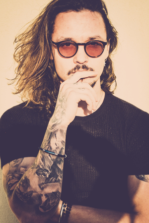 beautiful man with long black hair look at you thinking about it. Fashion model concept with vintage filter and warm tones. Black t-shirts and eyeglasses, trendy look and tattoesの写真素材