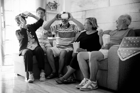 Aged senior group of grandfathers and teen at home trying goggles headset during a party in the afternoon with popcorn. Smiles and laugh for a funny timeの写真素材