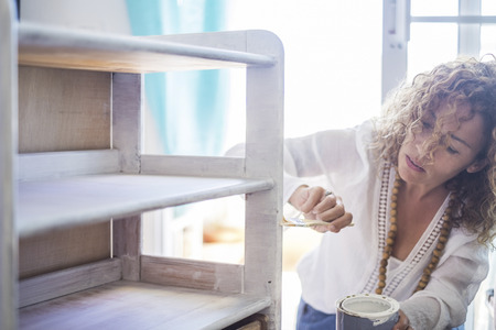 beautiful caucasian middle age independent woman paint and renew an old furniture at home with white shabby chic color. Do it yourself lifestyle for lonely and indipendent female. window light in the backgroundの写真素材