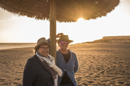 two senior women with hats on the beach under an umbrella. Sun Backlight of sunset. Leisure time in the summer in Tenerife.の写真素材
