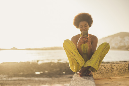beautiful black race african model with nice hair use the smartphone sit down near the ocean and the beach in freedom leisure vacation activity. contact and connect using wifi with far friends or parentsの写真素材