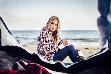 wild vacation alternative on the beach with a tent for a beautiful blonde female taking a mug with hot drink. Look at camera and rest. Alternative life concept.の写真素材