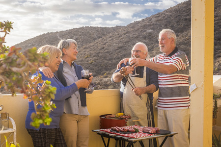 senior adult people group in leisure activity doing barbeque bbq on the rooftop terrace at home with mountain view. meal and wine for two men and two women having fun together under the sunlight in vacationの写真素材