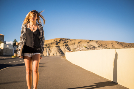 beautiful alternative rebel young woman 25 years old walking strong and independent with a skateboard on a road hill in tenerife during a sunny day. mountains in background.の写真素材