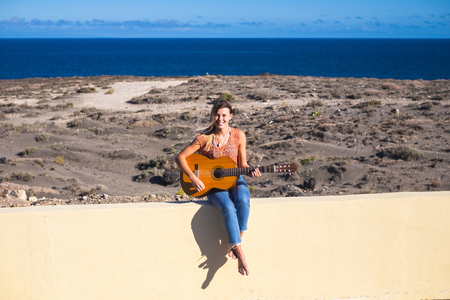 rebel and vacation alternative travel concept with beautiful caucasian blonde playing music with guitar outdoor near the ocean coast in tenerifeの写真素材