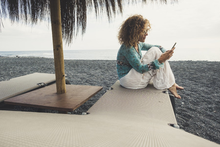 single nice middle age caucasian female at the beach sit down on the seats with ocean and sand around her. checking the phone to see internet and emails and work out of the office. stay connected conceptの写真素材
