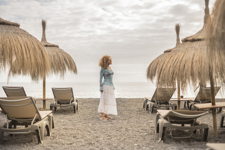centered position beautiful caucasian middle age woman freedom in outdoor leisure activity at the beach with ocean and clouds in the sky. seats and umbrellas spot and fashion dress with nice curtly hairの写真素材