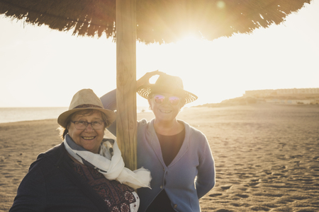 Couple of female seniors aged at the beach during the sunset in Tenerife. Leisure concept for 60s women with hats and backlight. Warm filter.の写真素材