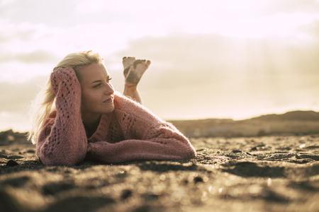 beautiful free model lay down on the sand at the beach enjoying an amazing relaxed golden sunset outdoor leisure activity. nice blonde girl young with nude and sandy feet look at his side. sexy and caucasian peopleの写真素材