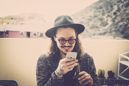 long hair nice young man read and check the phone on the roofteop terrace outdoor. black hat and eyeglasses and tattoo on hand. fashion model sith nice smile. modern hipster conceptの写真素材