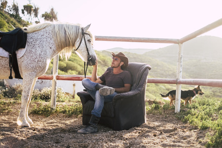 caucasian man enjoy his horse resting on a seats outdoor in the countryside. alternative lifestyle in contact with animals and nature. dog in the backgroundの写真素材
