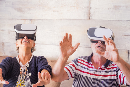 adult couple play and try modern goggled headset at home. hands on hands together with fun and fear. technology for seniorsの写真素材
