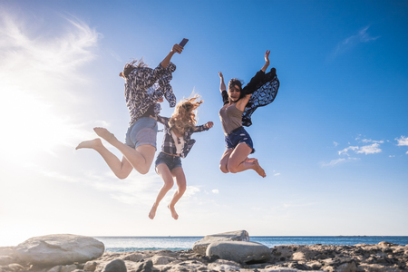 Group of three beautiful nice young woman jumping for fun and happiness, independent life together in friendship. Tenerife stone beach in a sunny day, enjoy the time.の写真素材