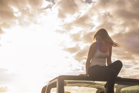 freedom and vacation concept for beautiful blonde caucasian young woman sitting down on the roof of a off road black car. Backlight and sunlight in warm tonesの写真素材