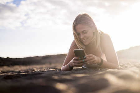 blonde young woman in vacation on the beach check the mobile smart phone to connect with friends. Lay down on the sand with sun backlightの写真素材