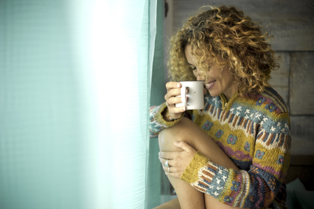 beautiful lonely caucasian woman with curly hair and smile white enjoy her loneliness drinking a tea or coffee at home in front of the window. sit down and hug herself. winter indoor conceptの写真素材