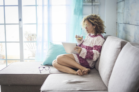 beautiful relaxed lady at home witting on the sofa and drinking a healhty water and lemon beverage while work with a tablet connected to internet to manage on line office and shop. alternative lifestyleの写真素材