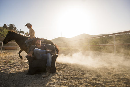 couple of beautiful man and woman together. she rides a horse making dust with speed and he stays sit down on the chair in the middle of the trackの写真素材