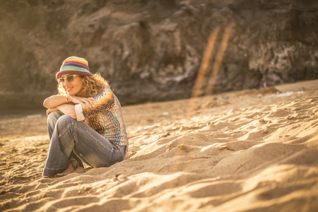 pretty blonde young woman sit down on the sand at the beach relaxing and enjoying sunset and sunny day of vacation. stay well in a natural place and outdoor leisure activity concept. colored clothesの写真素材