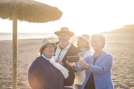 elderly seior group of friends with a 40s woman having phone at the beach with a mobile phone. Sunlight in backlight while they take a selfie.の写真素材