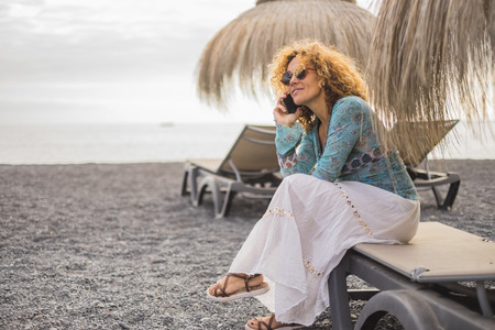 nice and smile beautiful middle age woman model 40 years old european with golden curly hair speaking at the phone with friends or parents like son or husband. sit down at the beach outdoor leisure activity with mobile internetの写真素材