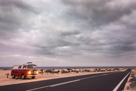 Red old van with lights on parked near the ocean. Long road across the desert of Fuerteventura Corralejo.の写真素材