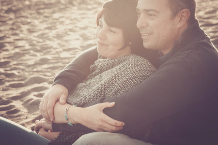 nice beautiful middle age 40 years old couple man and woman caucasian hugged and stay together on the beach sitting in the sand and enjoying a golde amazing sunset for nice leisure activity outdoor in the natureの写真素材