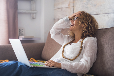 smile and laughing caucasian beautiful middle age woman sit down on the sofa at home with a colored keyboard laptop. working happy independent concept for nice modelの写真素材