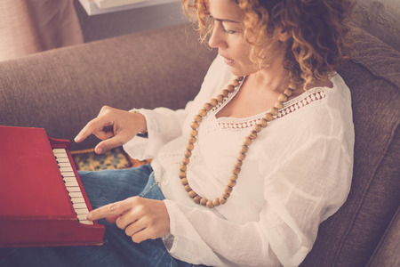 beautiful caucasian curly hair model middle age woman at home playing a little piano toy. artist at work sitting on the sofa. lonely female study music alone for hobby or workの写真素材
