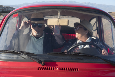 Elderly couple travelling inside a red car, having fun laughing with two hats. Vacation time and life conceptの写真素材