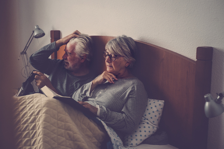 caucasian senior adult couple stay and rest on the bed. the wman read a book the man play with goggled headset technology. beautiful model and light from window.の写真素材