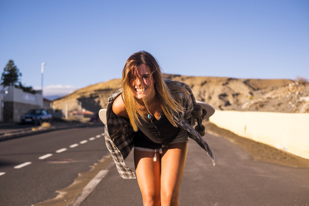 cute model beautiful blonde long hair smile to you in camera walking on a long road to travel with a skateboard behind. mountains background and sunlight day of vacation in tenerifeの写真素材