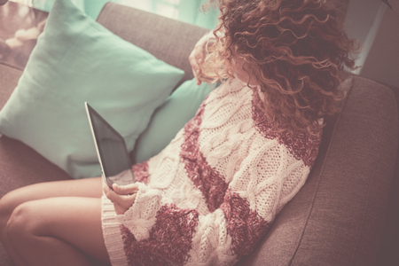 lonely lady at home with curly brown hair watching technology tablet connected to internet to check social media or work or movie. wifi and homeworking concept. woman sitting on sofa in leisure activity indoorの写真素材