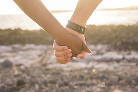 love concept with black and white hand touching and taking in front of the ocean during a golden sunset. warm light and yellow tones to represent true relationship with different races peopleの写真素材
