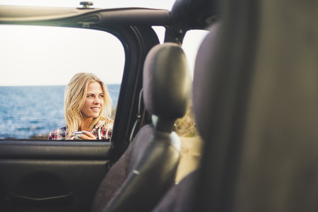 traveler blonde long hair girl take a mug with hot drink coffee or tea outside an open off road car. Ocean background and nice smile. Indipendent travel conceptの写真素材