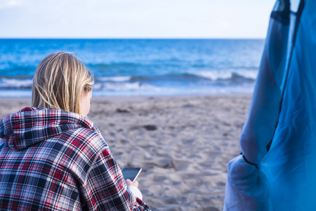 blonde beautiful woman sit down in spring on the beach out of the tent in free camping checking the phone for messagesの写真素材