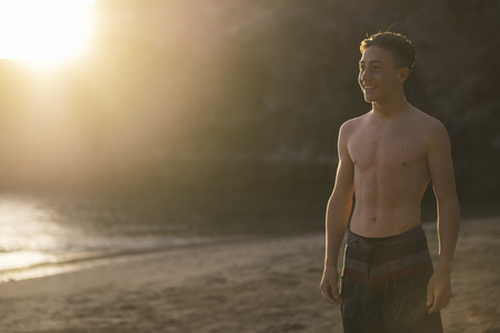 nice beautiful caucasian teenager with muscles at 15 years old standing on the beach looking the horizon and enjoying the golden light sunset in the background.の写真素材