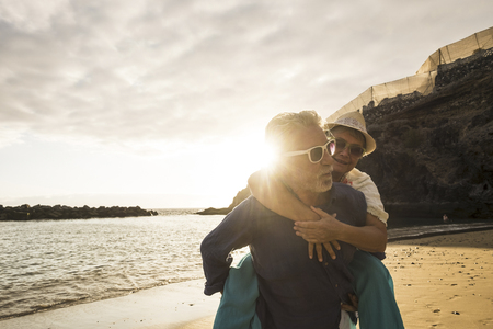 Happy senior adult caucasian couple in love. the man carry on his back the beautiful woman and both smiles. affective and together forever life concept in vacation time ith sunset backlight and ocean wavesの写真素材