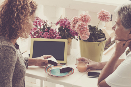 outdoor leisure activity talling storie for two caucasian women outdoor on the terrace, cake, fruit juice and tablet with internet and mobile phone for a nice afternoonの写真素材