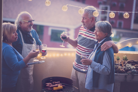 group of friends seniors having a bbq barbeque together on the rooftop with beautiful view on other buildings. Bulb lights in the terrace and smile having fun. two couples lesiure eldery concept. vintage filter.の写真素材