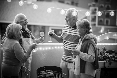 senior adult people group in leisure activity doing barbeque bbq on the rooftop terrace at home with mountain view. meal and wine for two men and two women having fun together under the sunlight in vacationの写真素材