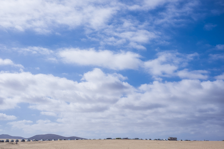 A lot of vehcles parked in Fuerteventura near the desert of Corralejo. Sand and summer with mountains in background and beautiful blue sky aboveの写真素材