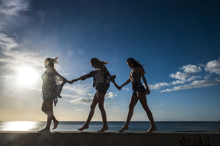 Three young women friends walk together on a wall near the beach in Tenerife. Enjoy the vacation under a blue sky with yellow sun. Hands by hands.の写真素材