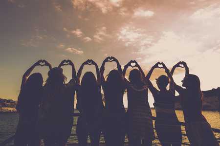 romantic concept for girls doing hearts with hands all together in relationship and friendship in front of a beautiful sunset and the ocean. golden tones for sweet image of group of people outdoorの写真素材